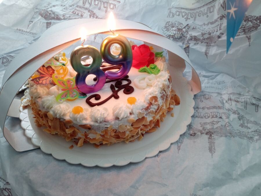 Tarta de cumpleaños, de nata, con una pinta de estar deliciosa. Sobre ella hay adornos comestibles con forma de mariposa y de flor. En el centro hay dos velas encendidas, con un 8 y un 9. 

89 años. 

Por la perspectiva de la foto parece que la llama de las velas está peligrosamente cerca de los cartones y el papel que envolvían la tarta, y de una guirnalda con forma de banderín, pero tranquilidad, no hemos quemado nada.